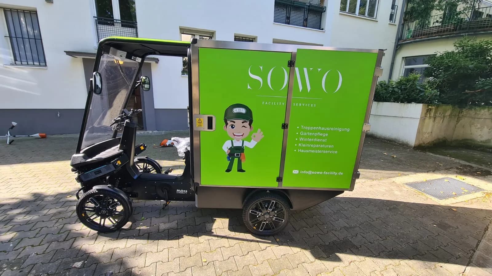 E-Lastenrad mit Fahrerkabine und Wetterschutz in der Seitenansicht vor einem Wohnhaus